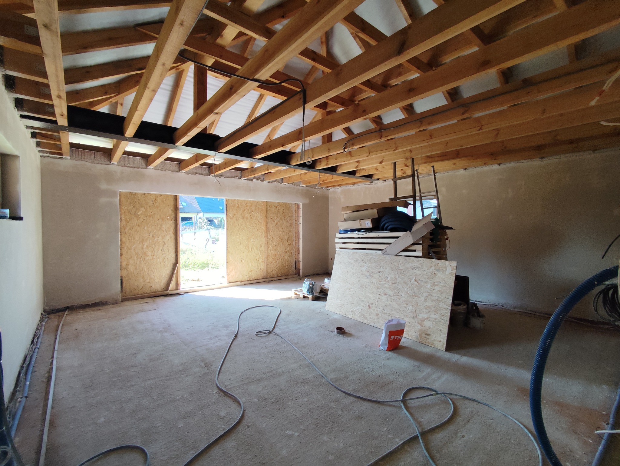 Wnętrze w trakcie prac wykończeniowych: surowy tynk na ścianach, drewniana konstrukcja dachu, przygotowane płyty OSB i widoczne przewody instalacyjne.
