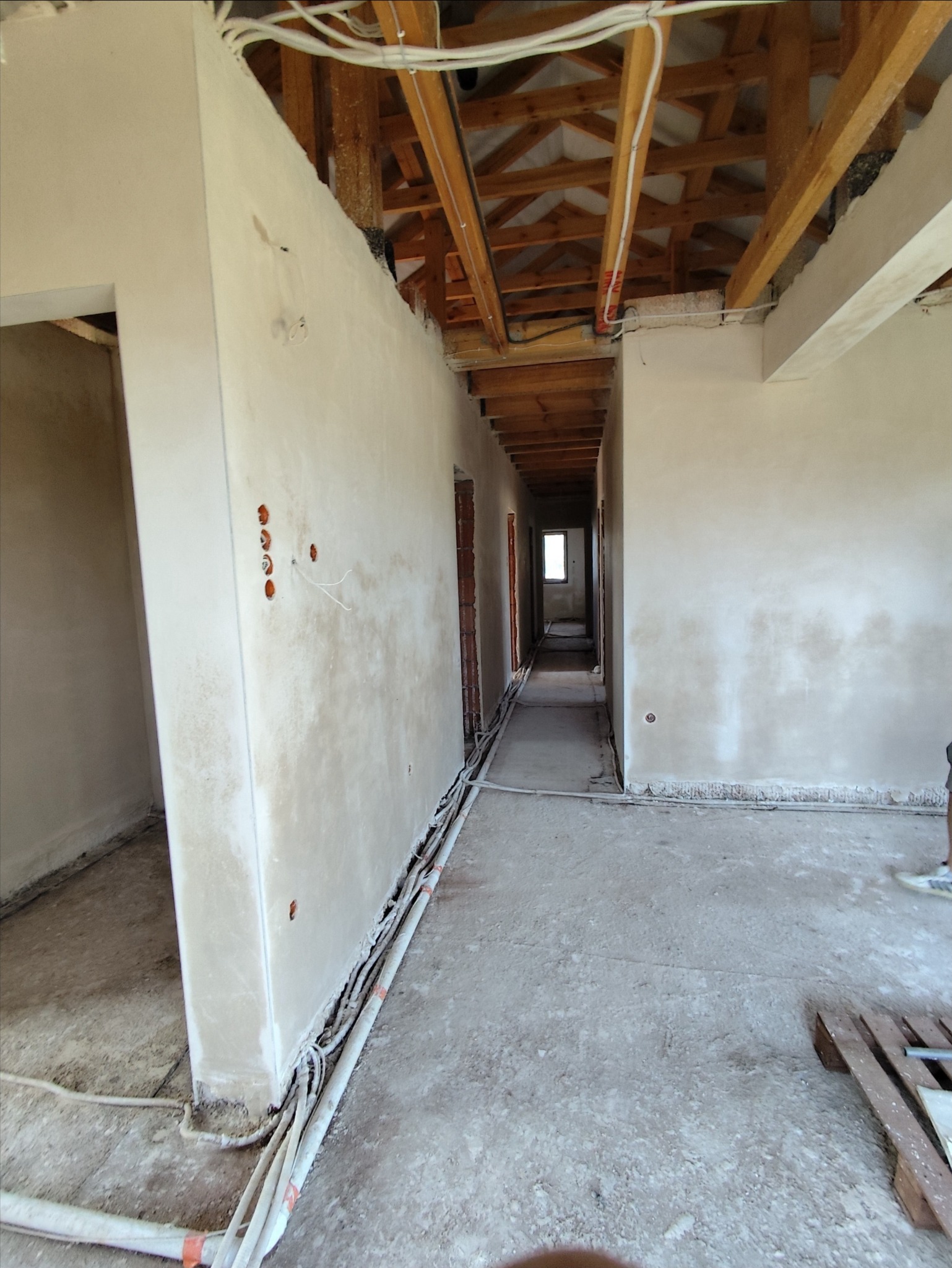 Korytarz w surowym stanie po nałożeniu tynków maszynowych, widoczne kable elektryczne wzdłuż ściany i drewniana konstrukcja dachu.
