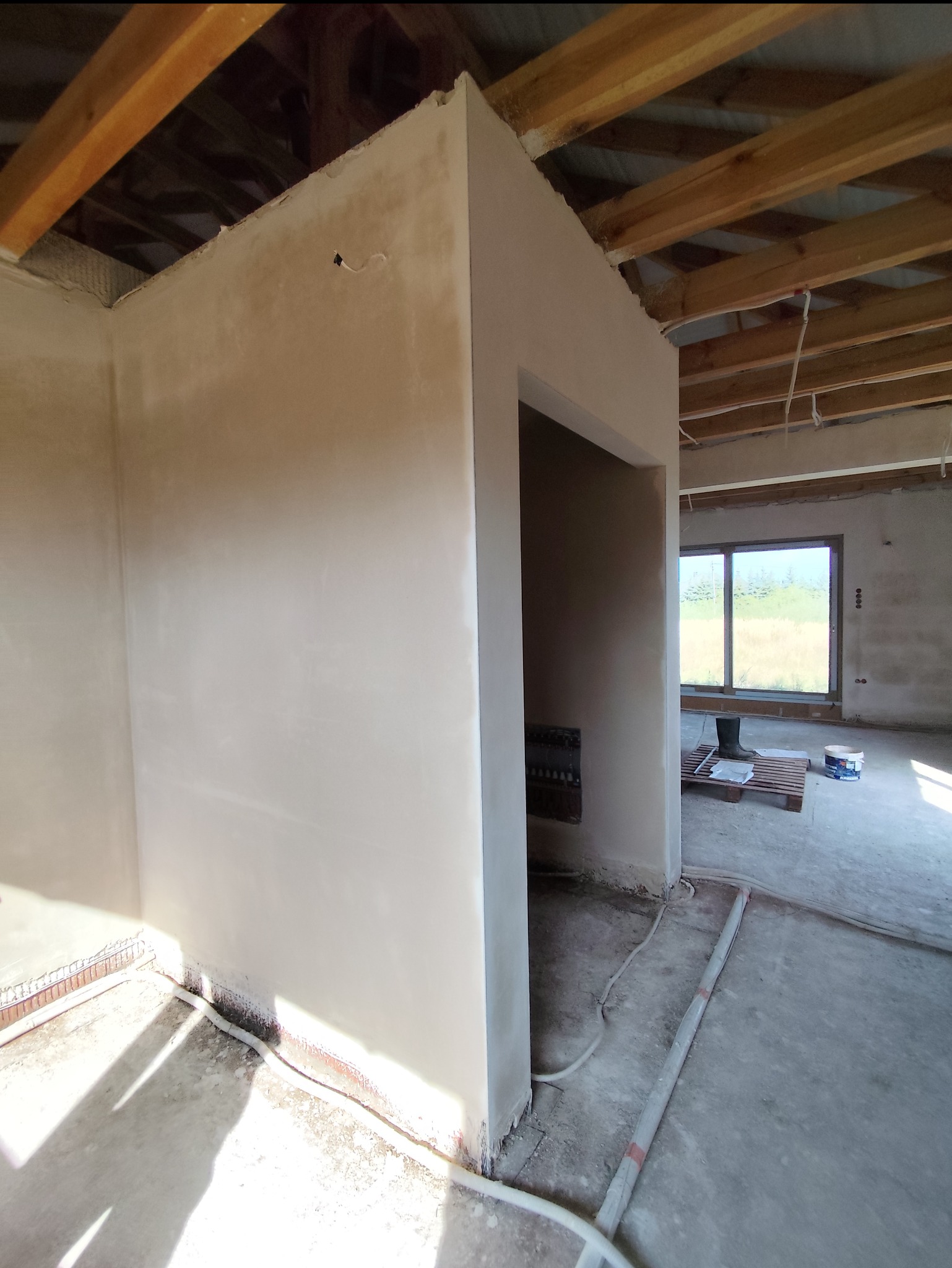 Wnętrze budynku w trakcie wykańczania, ściany pokryte świeżo nałożonym, jasnym tynkiem, widoczne drewniane elementy konstrukcji dachu i wystające przewody elektryczne.