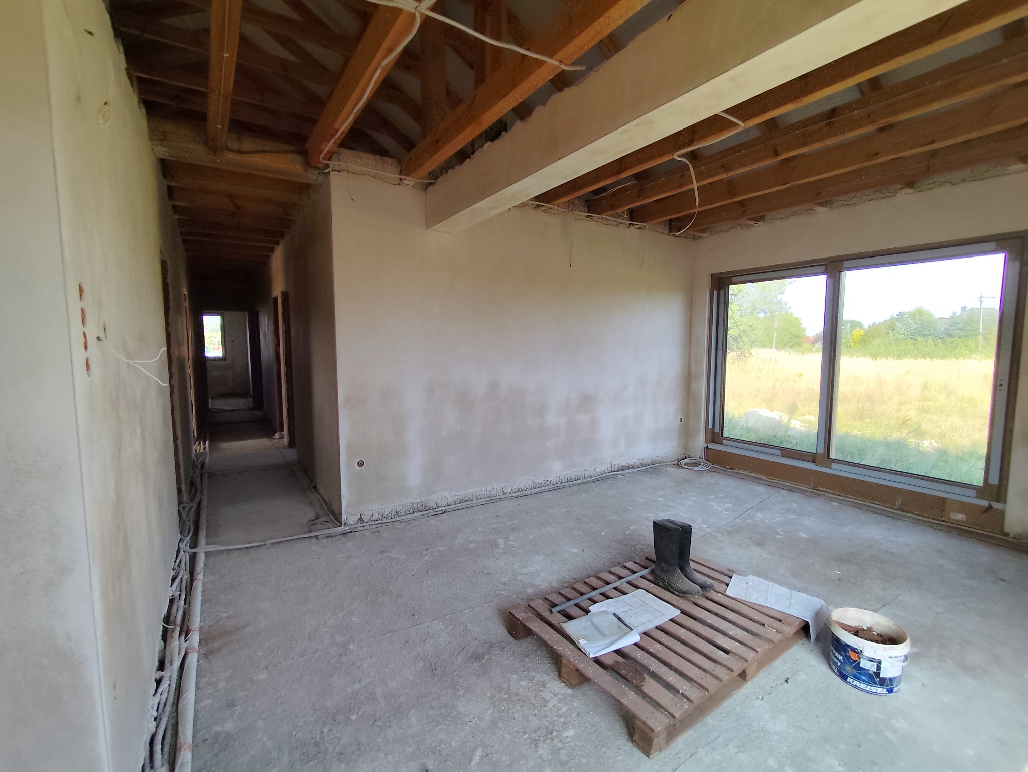 Wnętrze domu w trakcie tynkowania maszynowego, widoczne ściany pokryte świeżym tynkiem, drewniany strop, okno z widokiem na zewnątrz, paleta z roboczymi butami i dokumentacją oraz wiadro po farbie.