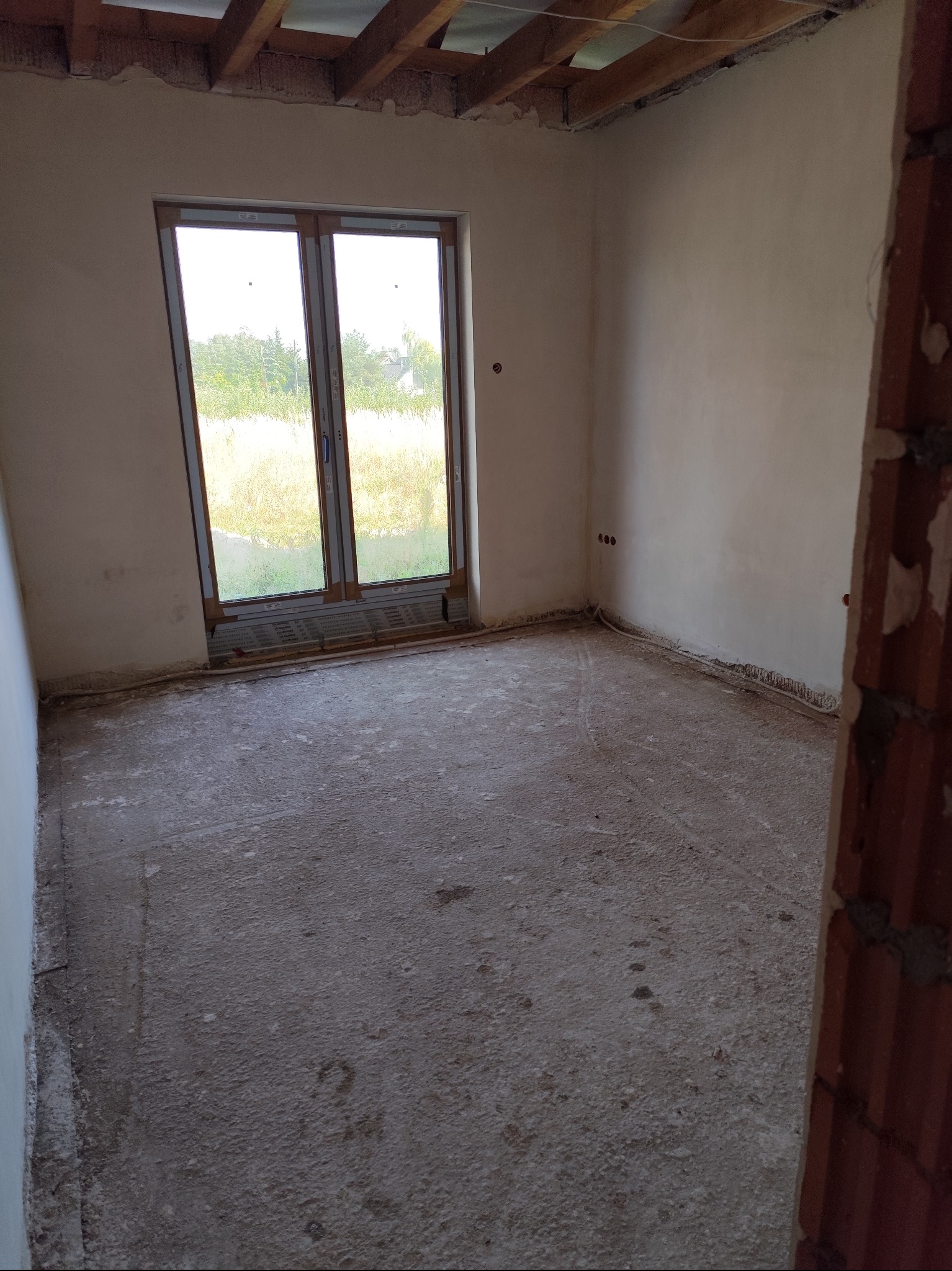 Surowe wnętrze pokoju z oknem, ściany pokryte świeżym, jasnym tynkiem, sufit z widocznymi belkami, podłoga betonowa w trakcie prac wykończeniowych.