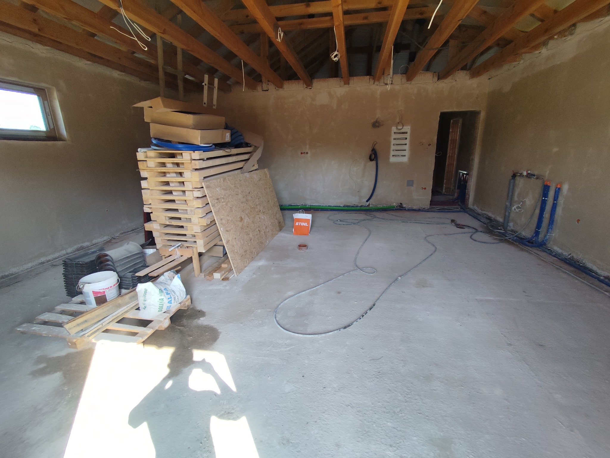 Wnętrze w trakcie prac wykończeniowych: surowe ściany po nałożeniu tynku, widoczne instalacje wodne i elektryczne, palety z materiałami budowlanymi i narzędziami, drewniana konstrukcja dachu.