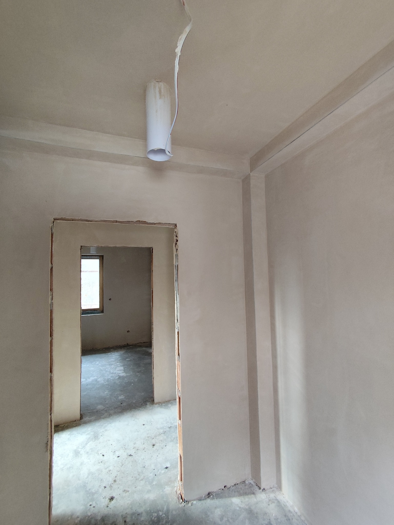 Wnętrze po tynkowaniu: gładkie, jasne ściany, widoczne narożniki i otwór drzwiowy. Minimalistyczne wykończenie, surowy stan przed dalszymi pracami. Wisząca lampa tymczasowa.