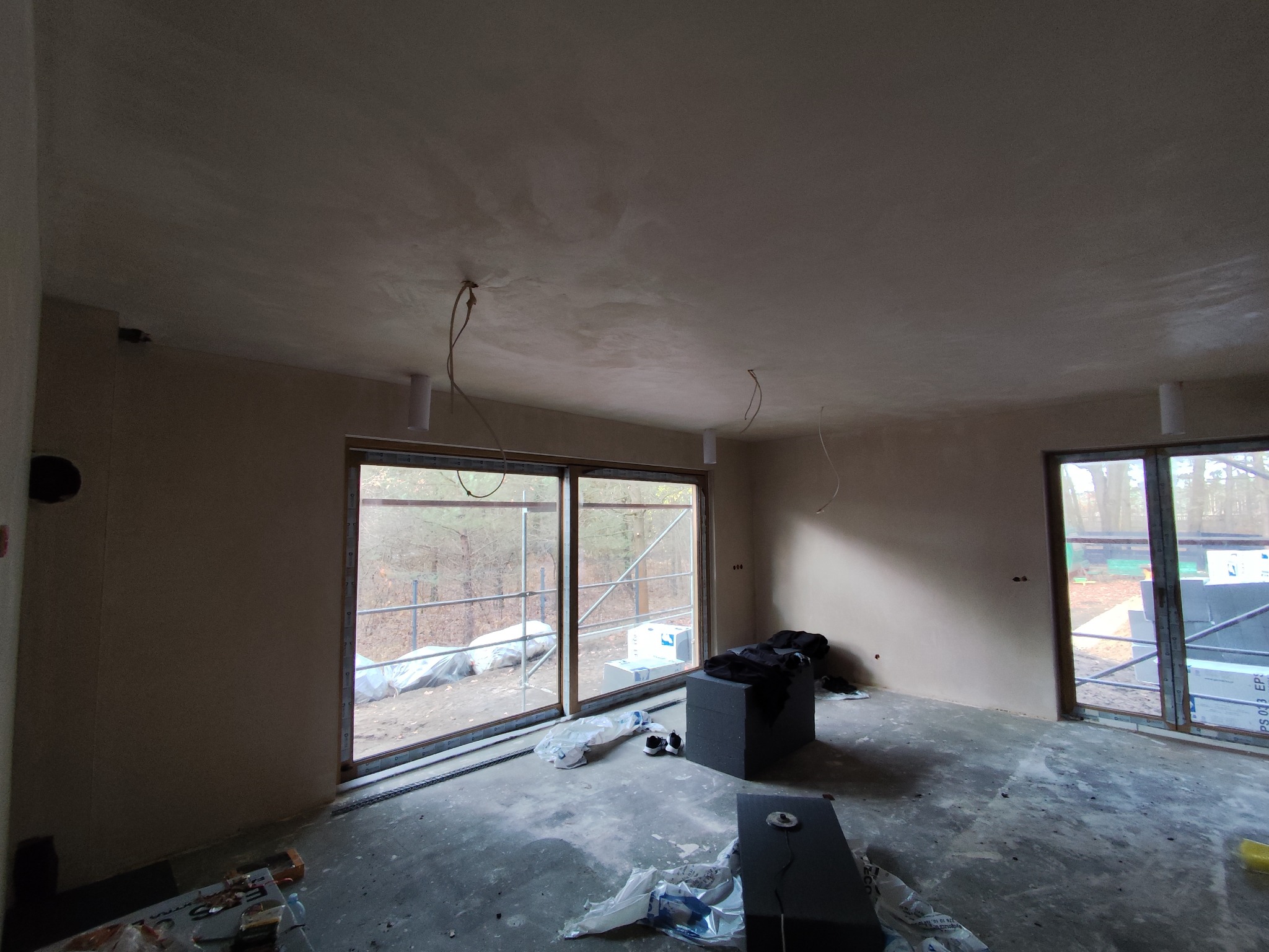 Wnętrze po tynkowaniu maszynowym: gładkie ściany, okna z widokiem na zewnątrz, widoczne przewody elektryczne, przygotowane do dalszych prac wykończeniowych.