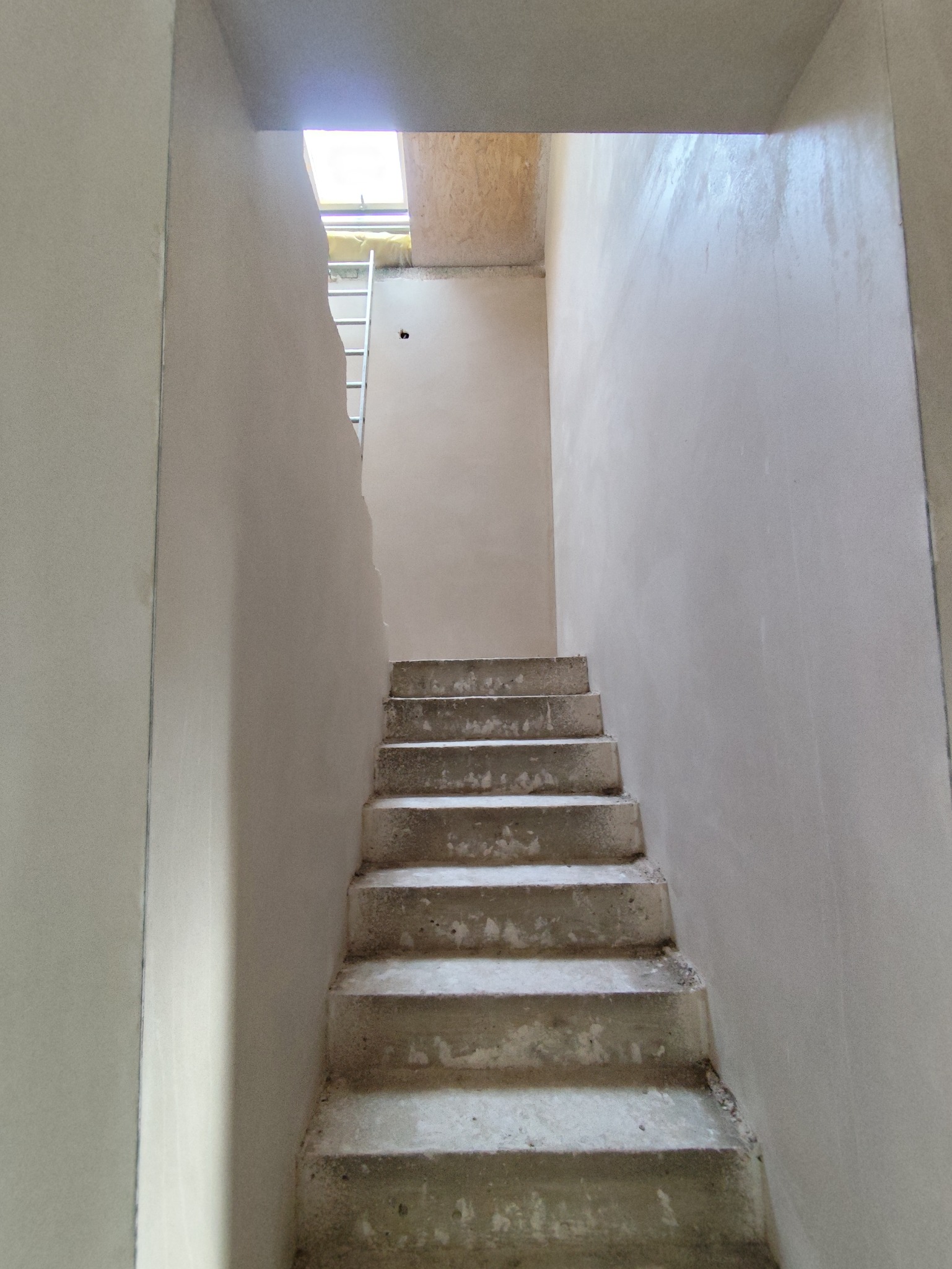 Wnętrze w trakcie prac wykończeniowych: surowe schody prowadzą w górę, ściany pokryte świeżym, jasnym tynkiem. Widoczny fragment drabiny i okno dachowe.