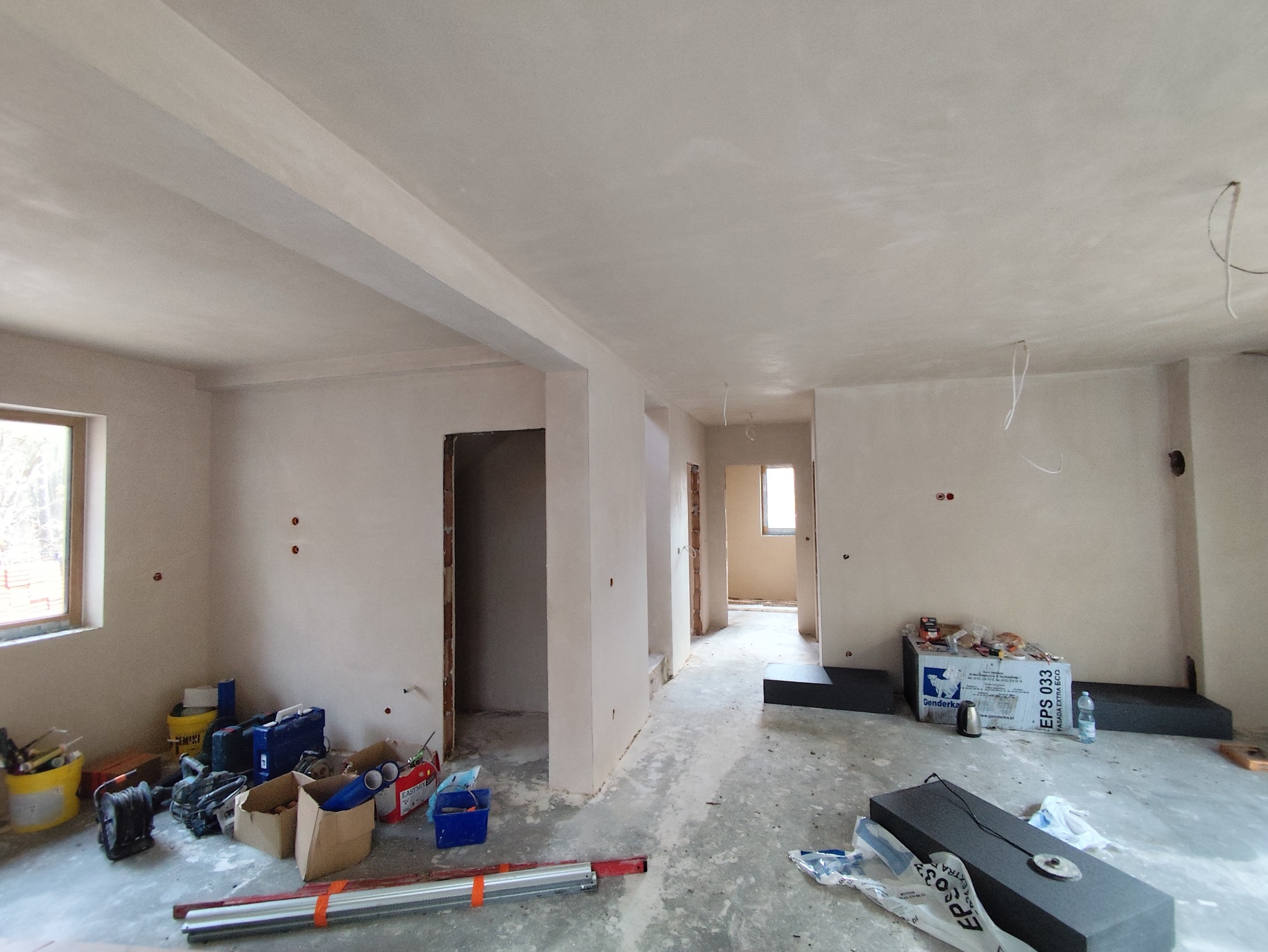 Wnętrze po tynkowaniu maszynowym: gładkie, jasne ściany, widoczne narzędzia i materiały budowlane, surowy stan wykończenia, otwory na gniazdka.