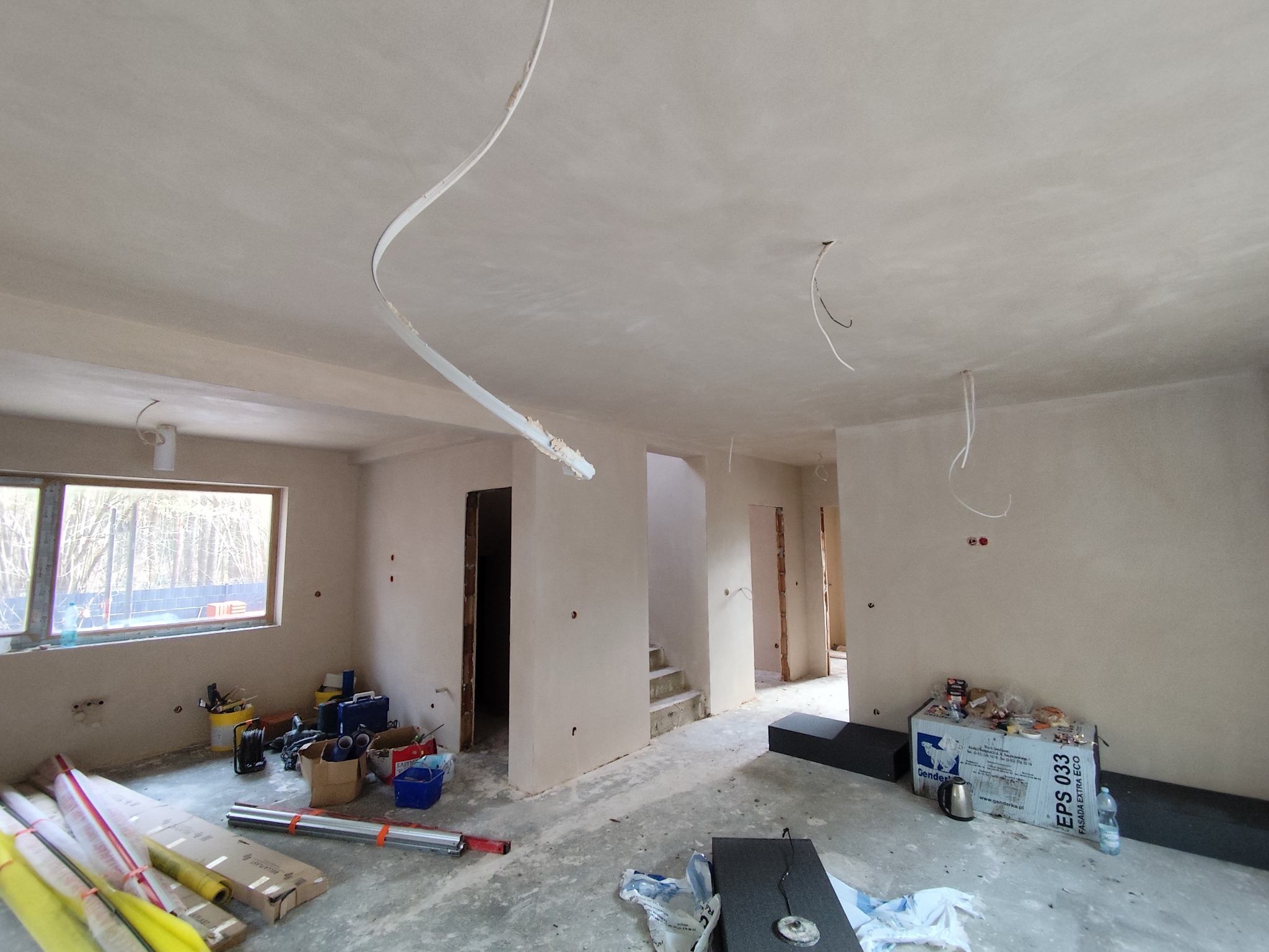 Wnętrze po tynkowaniu maszynowym: gładkie ściany, widoczne przewody elektryczne, okno z widokiem na las, narzędzia i materiały budowlane na podłodze. Jasne, przestronne pomieszczenie.