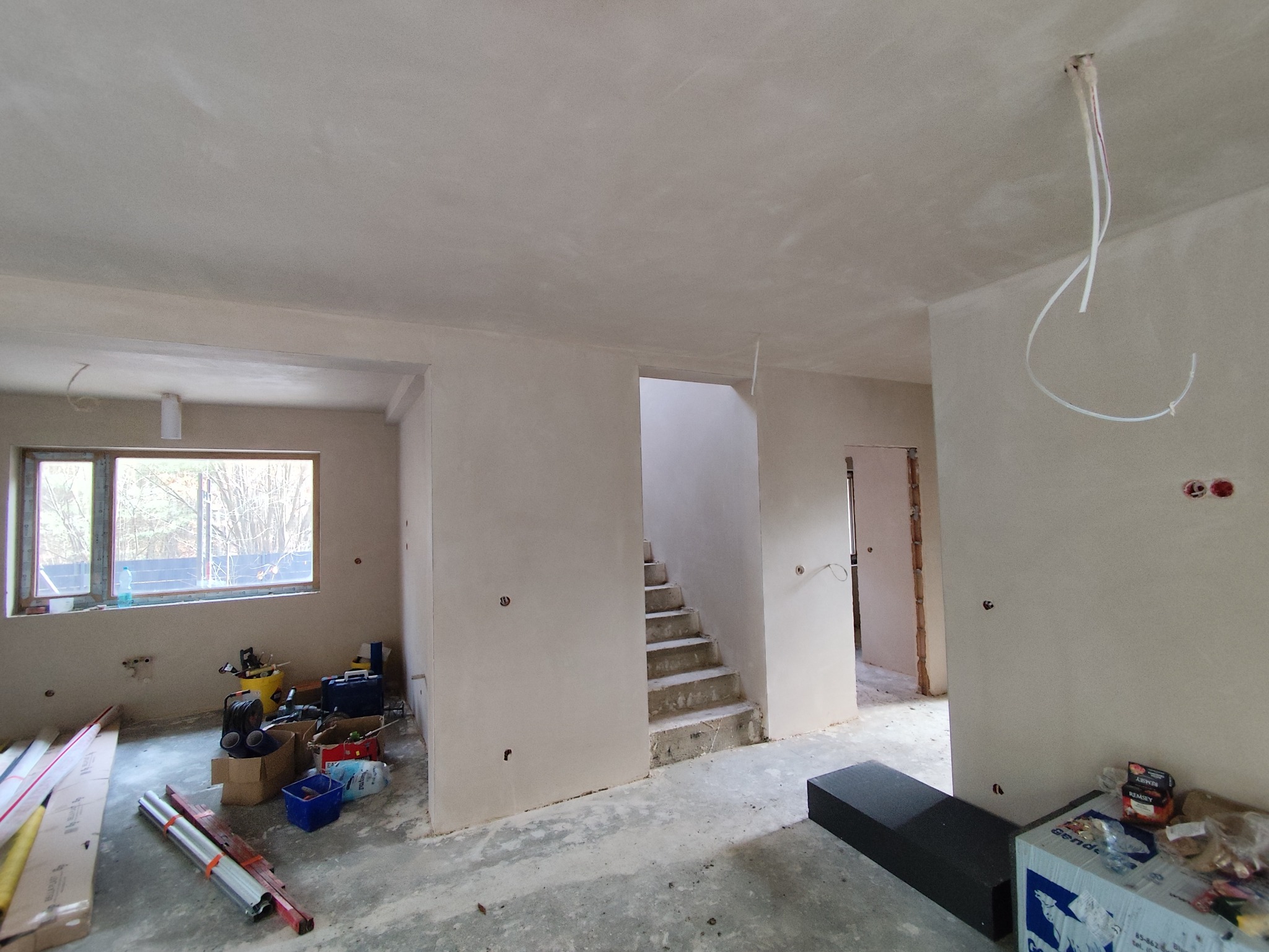 Wnętrze po tynkowaniu maszynowym: gładkie ściany, surowa klatka schodowa, widoczne instalacje elektryczne, okno z widokiem na zewnątrz, narzędzia i materiały budowlane.