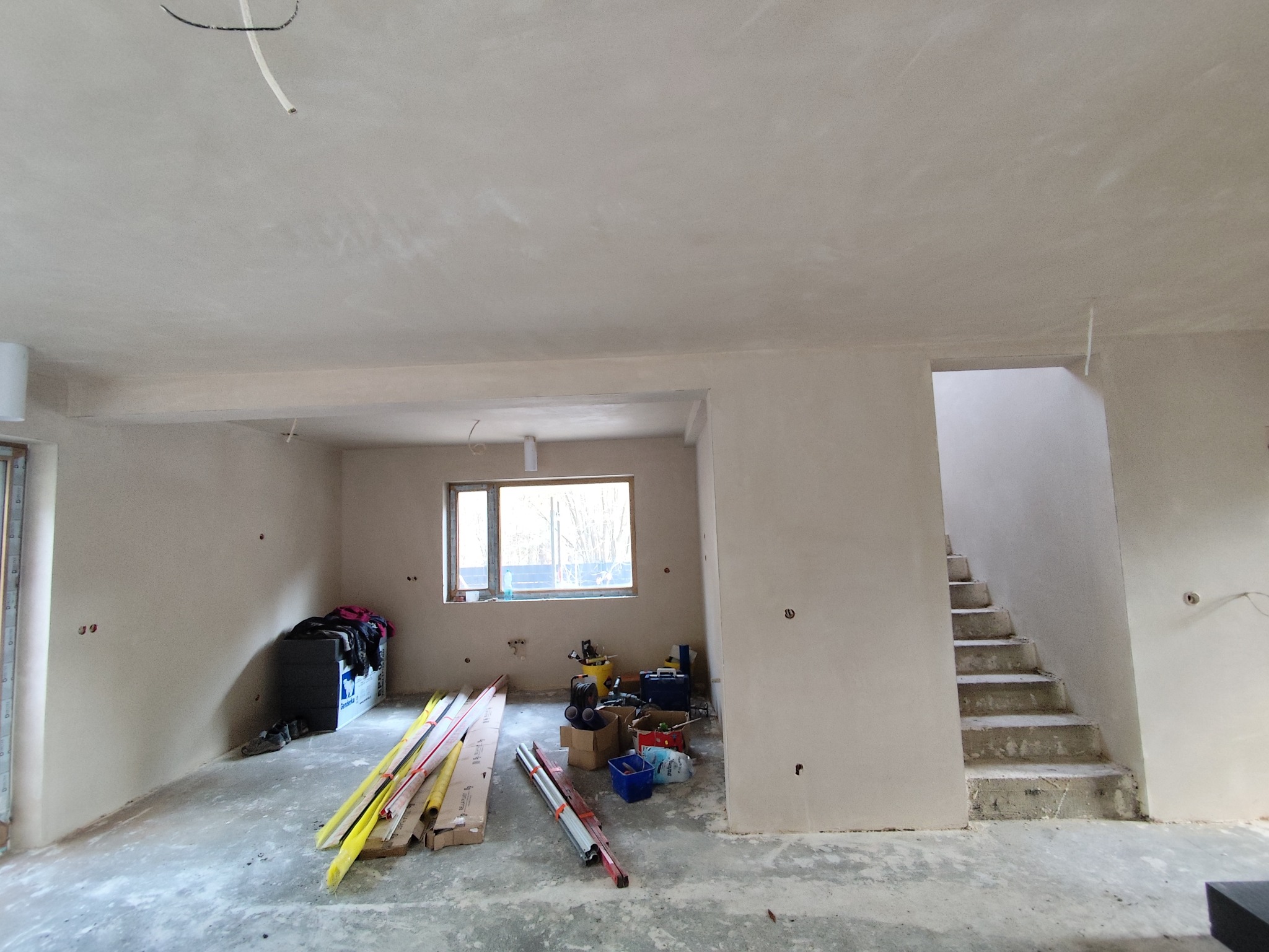 Wnętrze po tynkowaniu maszynowym: gładkie ściany, surowe schody, narzędzia i materiały budowlane. Widok na okno i przygotowane podłoże.