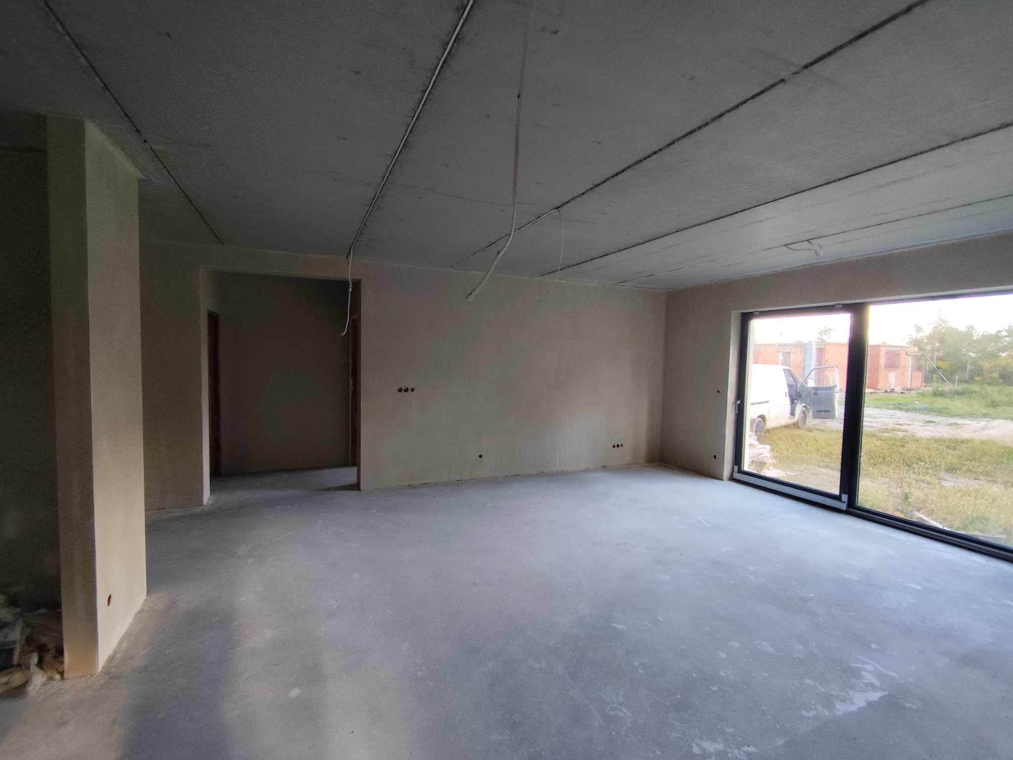 Surowe wnętrze po tynkowaniu maszynowym: gładkie ściany, betonowy sufit z instalacją, duże okno z widokiem na zewnątrz. Widoczny fragment podłogi i otwory drzwiowe.
