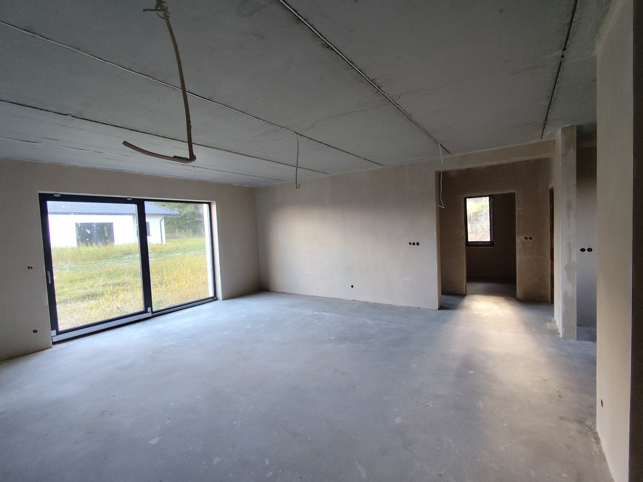 Wnętrze po tynkowaniu maszynowym: gładkie ściany i sufit w surowym stanie, widoczne przewody instalacyjne, duże okno przesuwne z widokiem na zewnątrz.