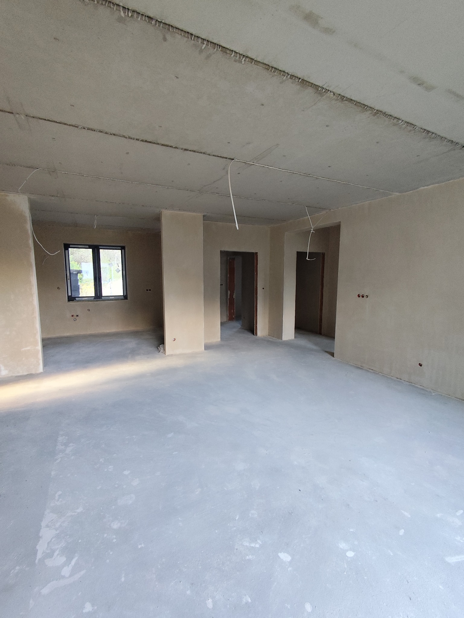 Wnętrze domu po tynkowaniu maszynowym: gładkie, szare ściany i podłoga, widoczne otwory drzwiowe i okno. Minimalistyczny, surowy stan wykończenia.