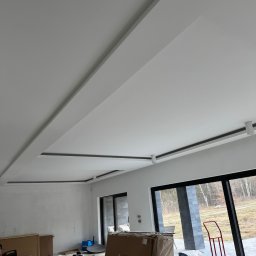 Silva budownictwo - Nowoczesny sufit podwieszany z oświetleniem punktowym w jasnym pomieszczeniu. Widoczne kartony, folia i wózek transportowy sugerują prace wykończeniowe.