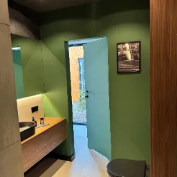 Nowoczesna łazienka w odcieniach zieleni z drewnianą szafką pod umywalką i czarną toaletą. Na ścianie wisi czarno-biała fotografia. Widok na ogród przez otwarte drzwi.