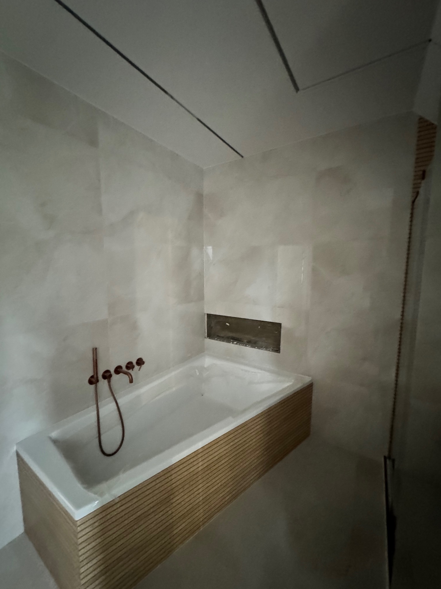 Nowoczesna łazienka z wanną obudowaną drewnem, kremowe płytki na ścianach i miedziana armatura. Wnęka w ścianie, minimalistyczny design.