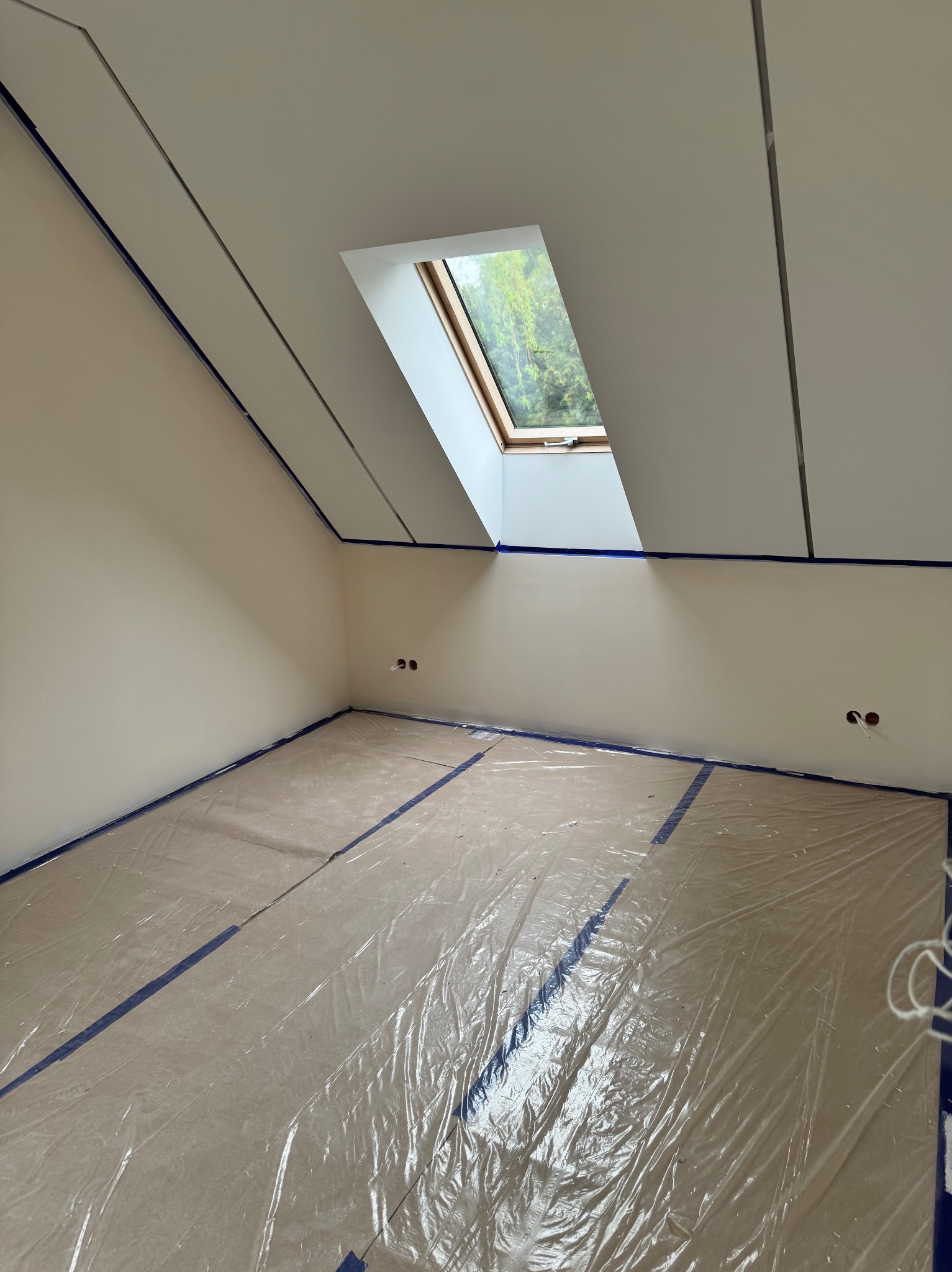 Pokój na poddaszu po szpachlowaniu, zabezpieczony folią malarską, z oknem dachowym i widocznymi gniazdkami elektrycznymi. Ściany w jasnym odcieniu beżu.