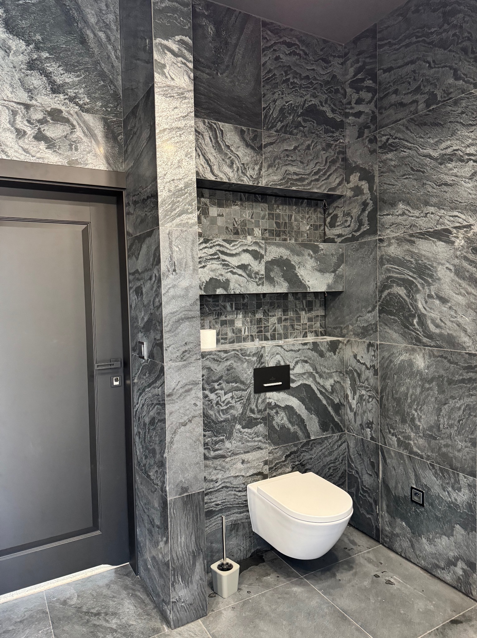 Elegancka łazienka w odcieniach szarości: ściany i podłoga wyłożone płytkami imitującymi marmur, wisząca toaleta i wnęki z mozaiką, tworzą spójną i nowoczesną przestrzeń.