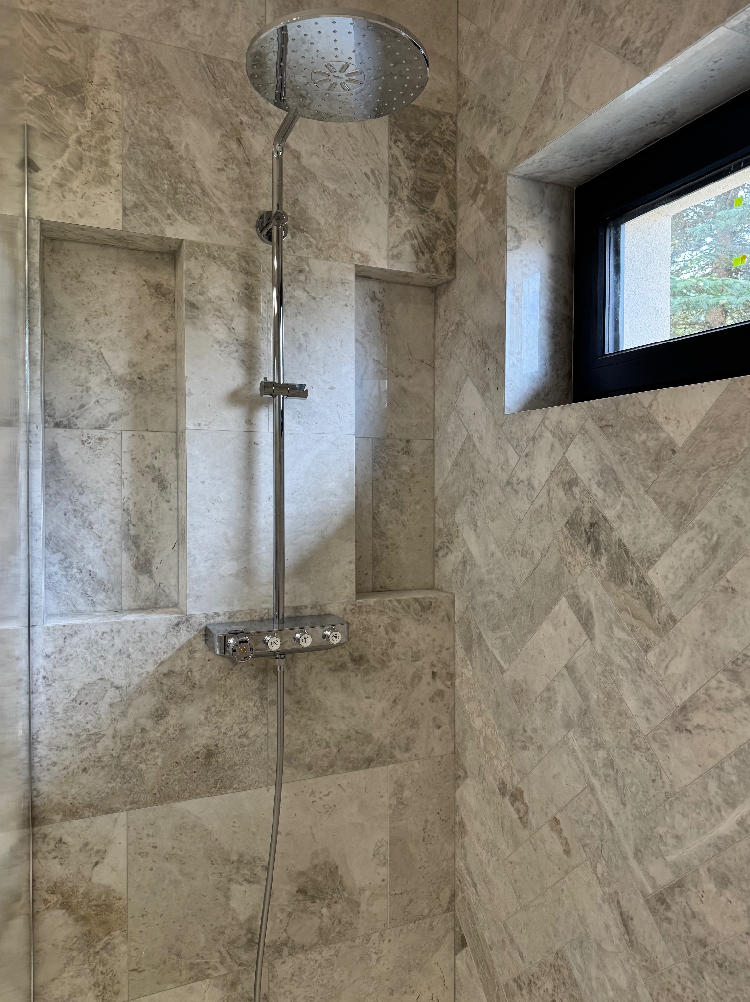 Wykończona kabina prysznicowa z armaturą w kolorze chromu i wnękami w ścianie wyłożonej marmurowymi płytkami ułożonymi w jodełkę, z oknem w tle.