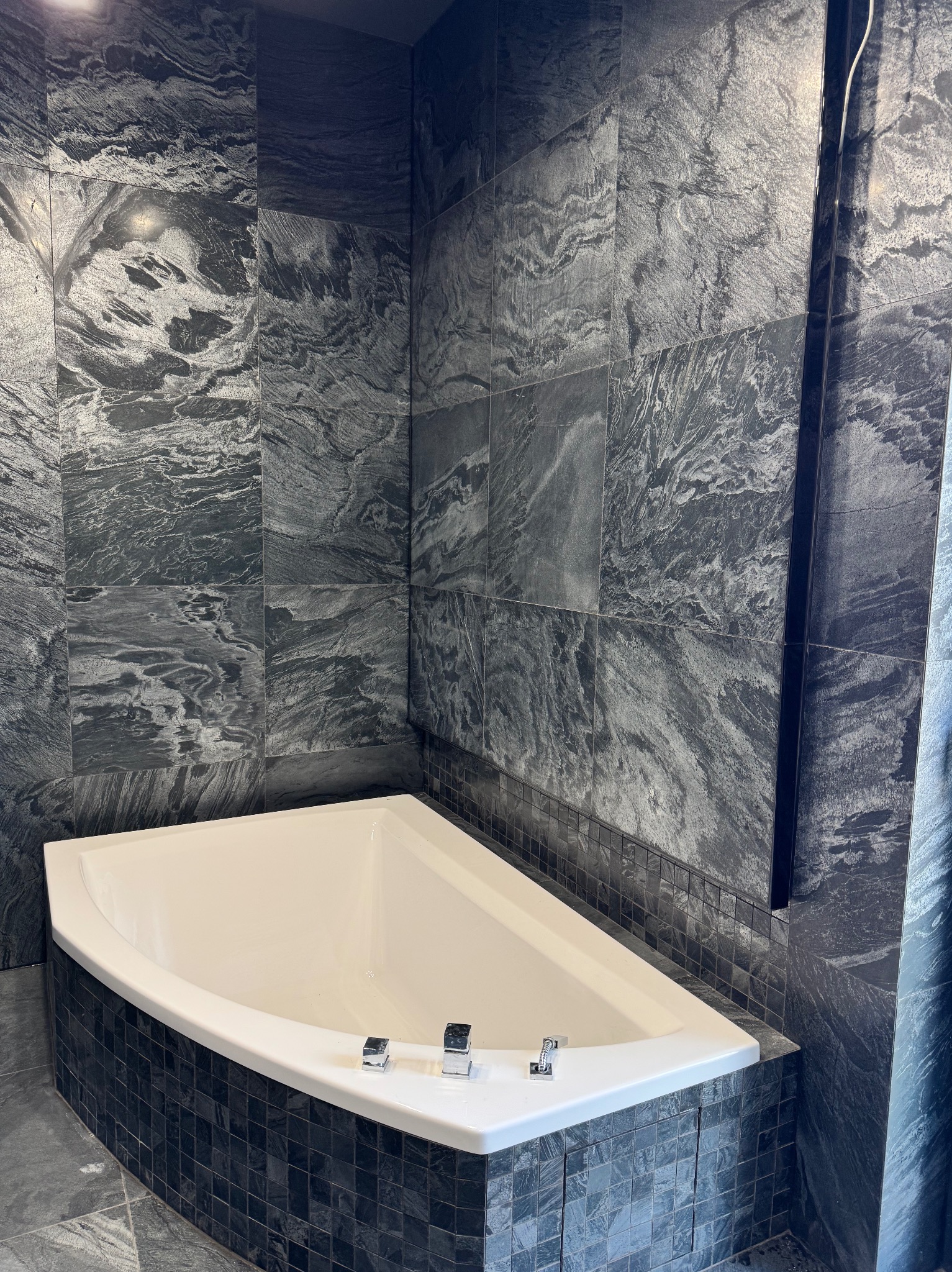 Wykończona łazienka z narożną wanną obudowaną mozaiką i ścianami wyłożonymi płytkami imitującymi marmur w odcieniach szarości. Nowoczesny design.