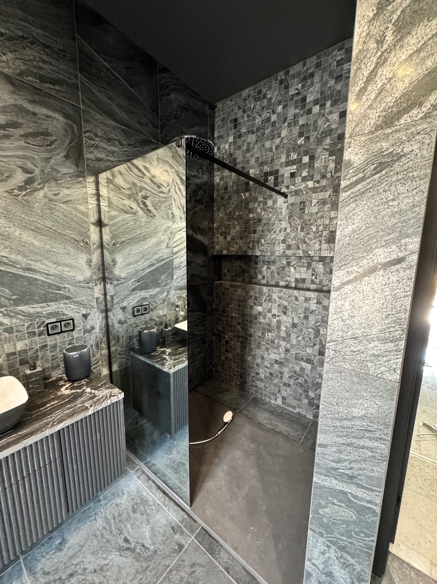 Nowoczesna łazienka w odcieniach szarości z kabiną typu walk-in, mozaiką i umywalką z szafką o ryflowanych frontach. Minimalistyczny design z akcentem na naturalny kamień.