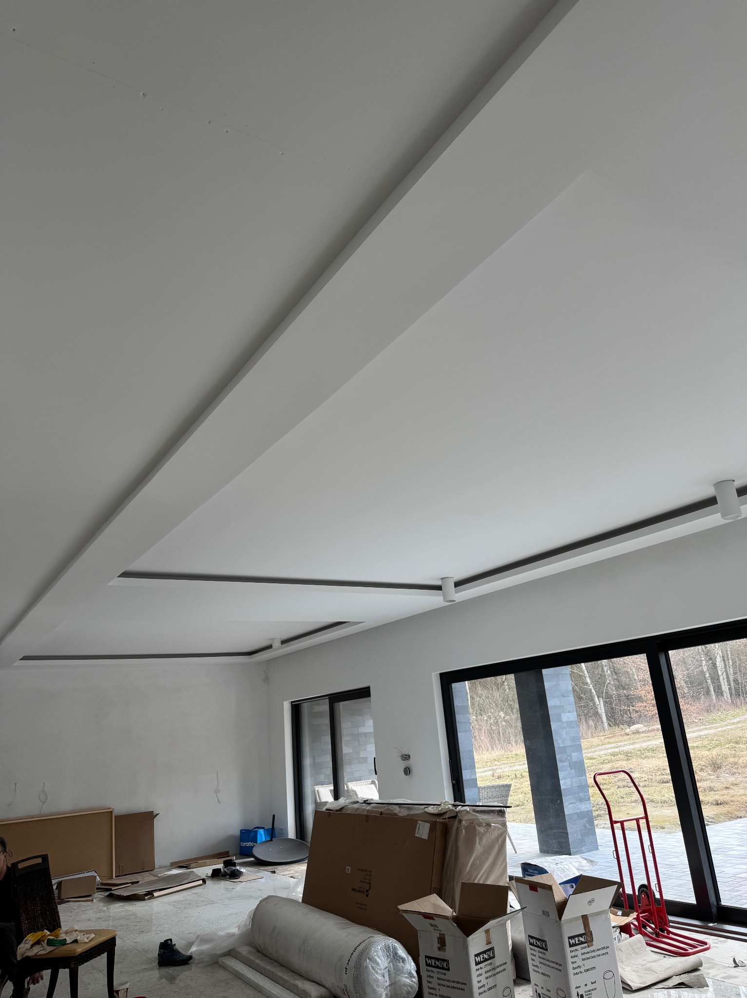 Nowoczesny sufit podwieszany z oświetleniem punktowym w jasnym pomieszczeniu. Widoczne kartony, folia i wózek transportowy sugerują prace wykończeniowe.