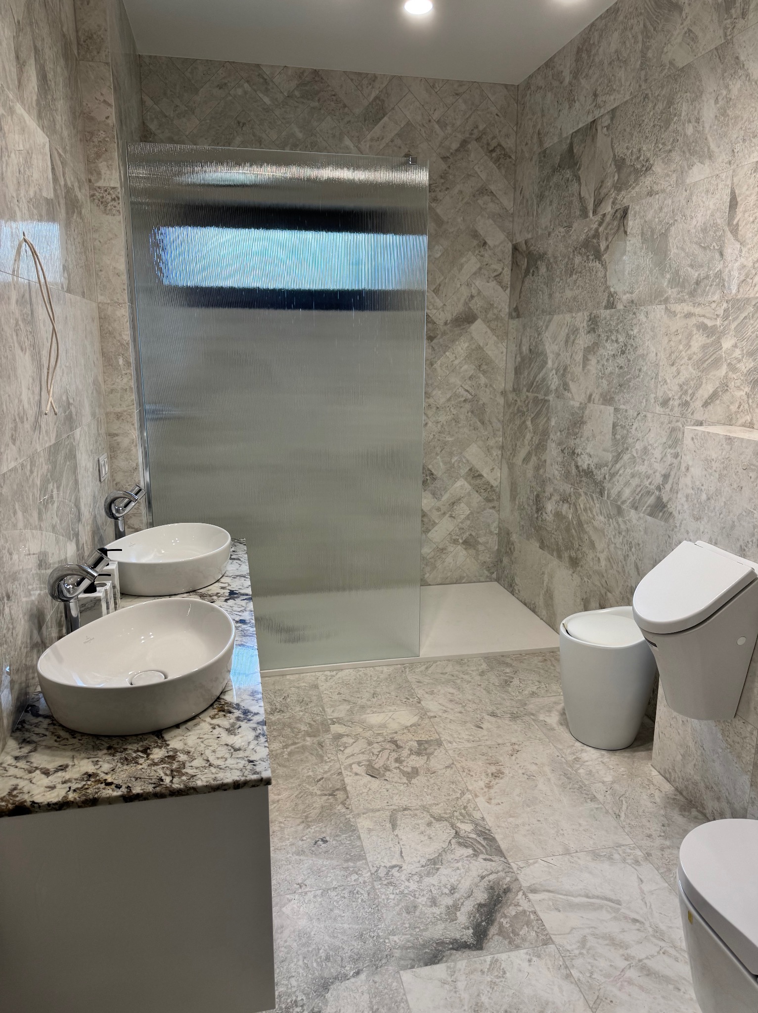 Wykończona łazienka z marmurowymi płytkami, kabiną prysznicową ze szkła ryflowanego i podwójną umywalką na blacie z kamienia. Nowoczesny design i stonowana kolorystyka.