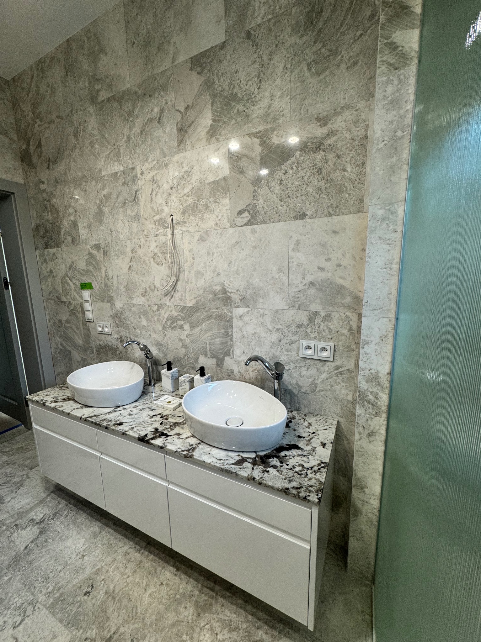 Nowoczesna łazienka: dwie białe umywalki nablatowe na szafce z blatem imitującym marmur. Ściany i podłoga wyłożone szarymi płytkami w marmurkowy wzór.