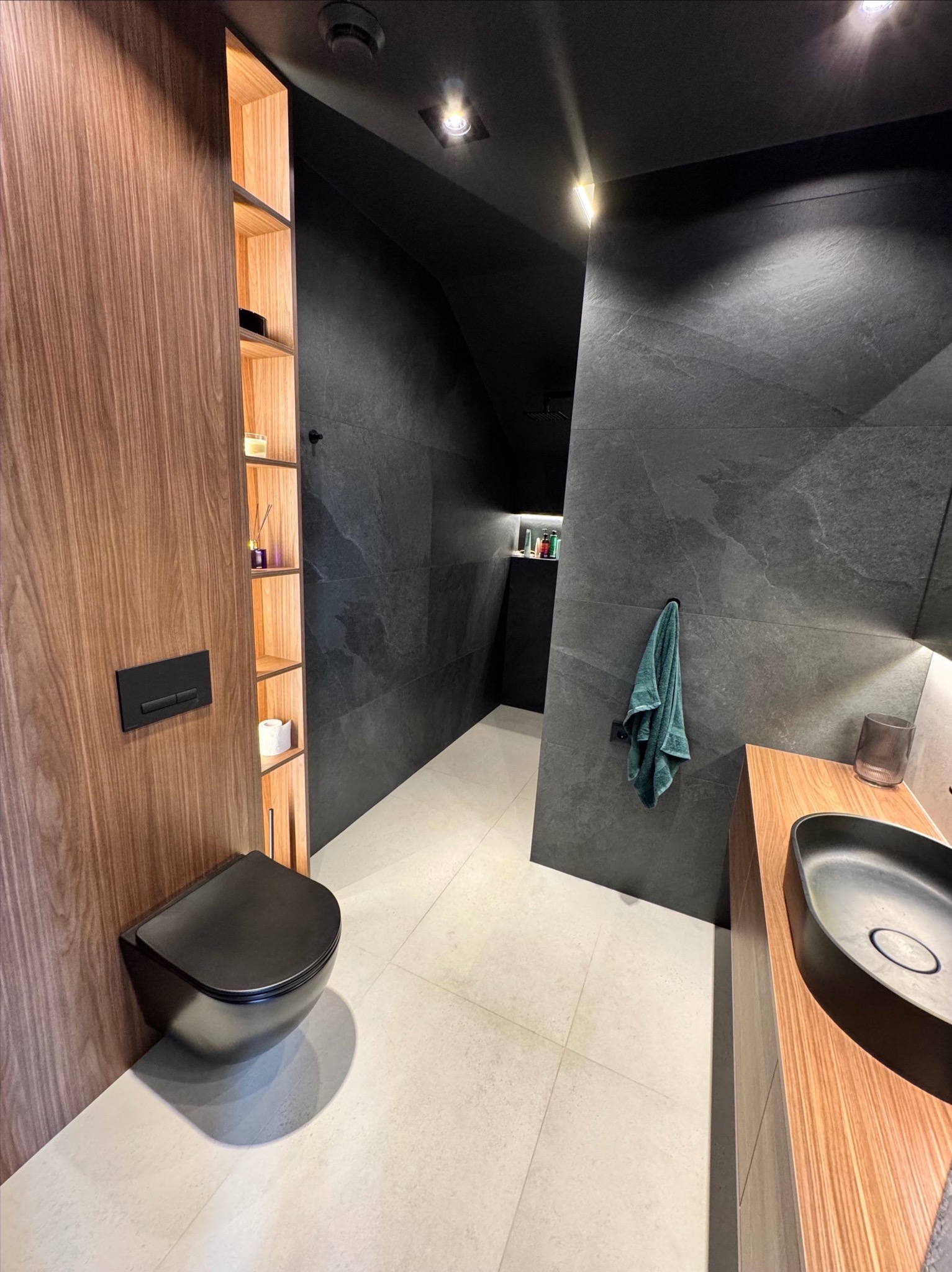 Nowoczesna łazienka w ciemnych tonacjach z drewnianymi akcentami. Uwagę zwraca podwieszana toaleta i umywalka w kolorze czarnym oraz oświetlenie podkreślające strukturę płytek.