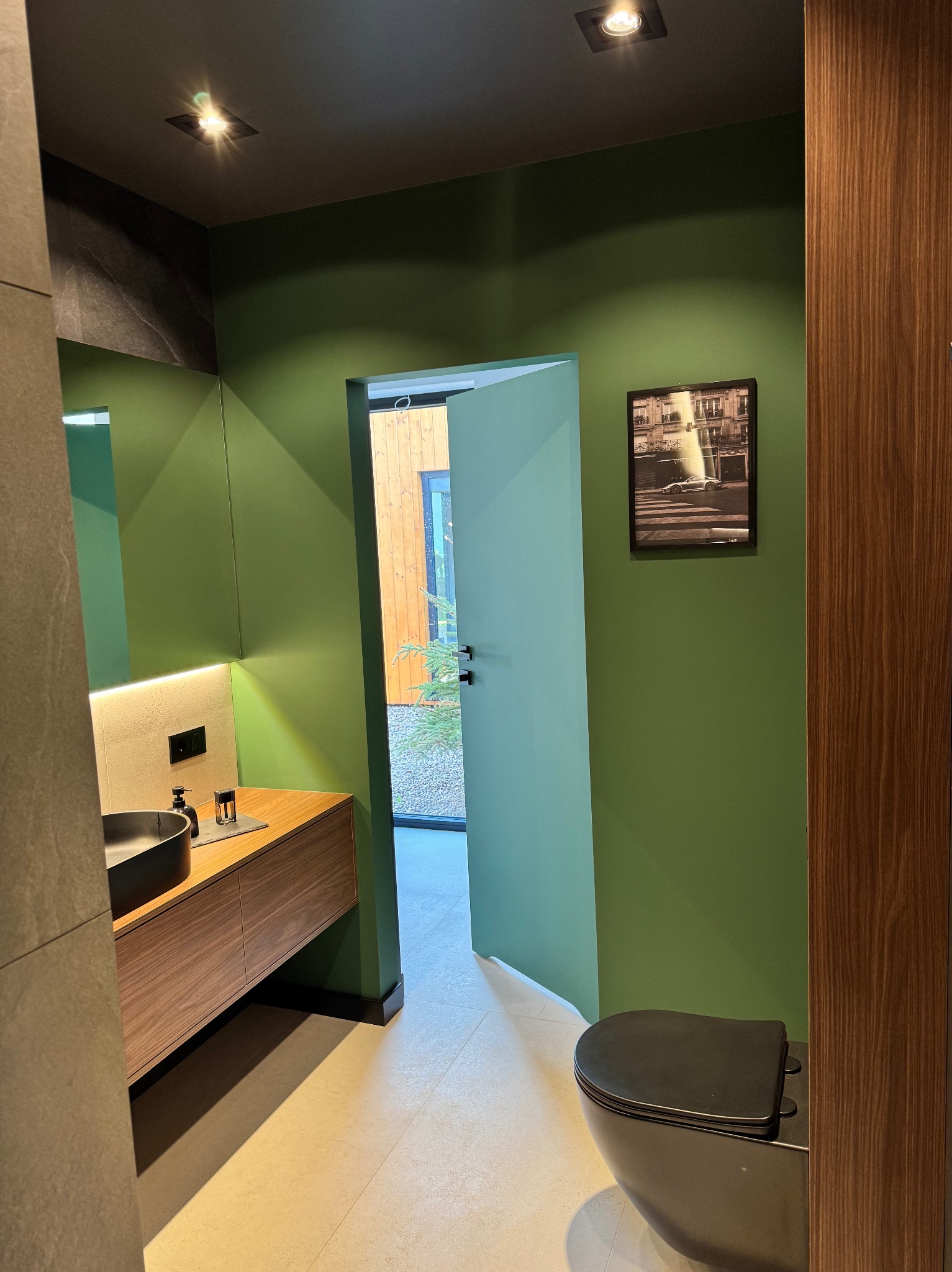 Nowoczesna łazienka w odcieniach zieleni z drewnianą szafką pod umywalką i czarną toaletą. Na ścianie wisi czarno-biała fotografia. Widok na ogród przez otwarte drzwi.