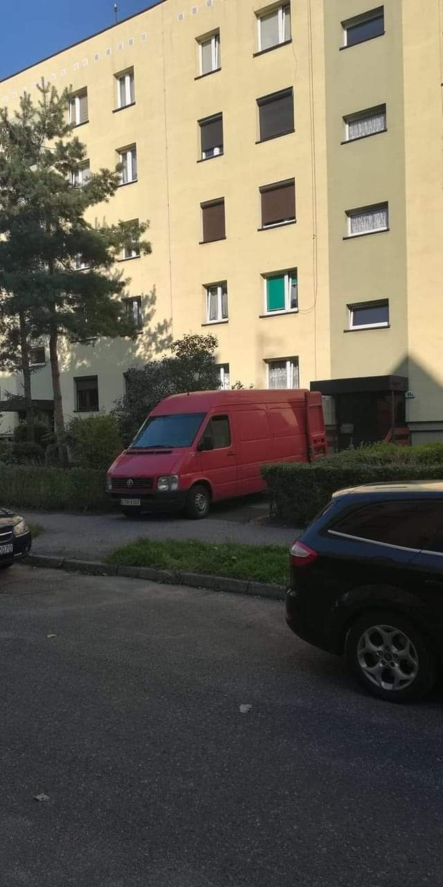 Czerwony bus dostawczy zaparkowany przed żółtym blokiem mieszkalnym z zieloną roletą w jednym oknie.