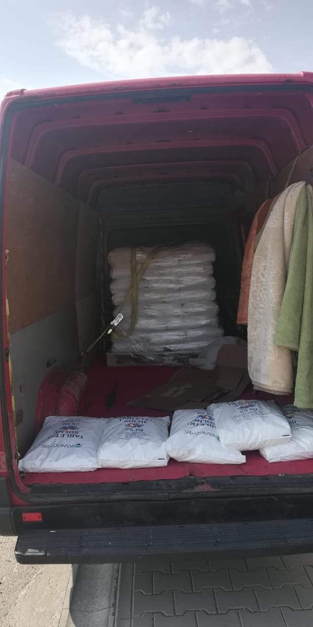 Wnętrze czerwonego busa załadowane paletą zafoliowanych worków i kilkoma workami z napisem 'TABLETKI SOLNE 25 KG', zabezpieczone pasami transportowymi, z wiszącymi ubraniami po prawej stronie.