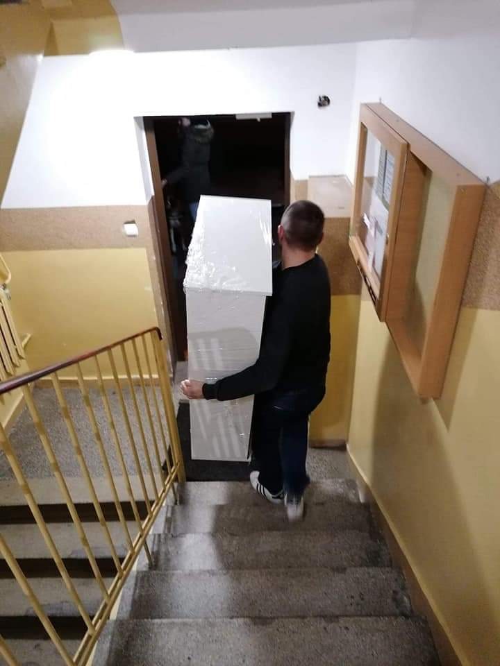 Mężczyzna w czarnej bluzie i dżinsach wnosi po schodach zapakowany w folię biały mebel, widok z tyłu, klatka schodowa z żółtymi ścianami i metalową barierką.