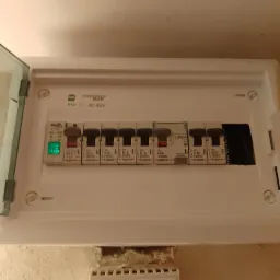 Otwarta skrzynka bezpiecznikowa z widocznymi bezpiecznikami Legrand o różnym amperażu, zamontowana na pomalowanej ścianie.