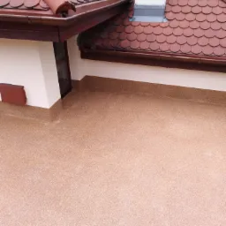 Wykończony taras na dachu z beżową, żywiczną nawierzchnią, widok z góry na krawędź dachu z czerwoną dachówką i rynną.