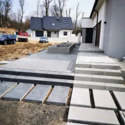 Nowoczesna posesja z szarymi płytami chodnikowymi ułożonymi wzdłuż ścieżki, obok widać przygotowane podłoże i luźno ułożone płyty, w tle dom z ciemną dachówką i samochody.