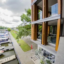 Widok z góry na nowoczesny apartamentowiec z drewnianymi okiennicami, przylegający do przystani jachtowej z licznymi łodziami, zieleń drzew i most w tle.