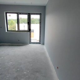 GMK URZEDOWSKI - Świeżo pomalowane na szaro wnętrze pokoju z oknem i drzwiami balkonowymi; widoczne ślady farby na podłodze.