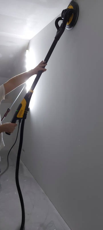 Szlifierka teleskopowa z odkurzaczem w dłoni osoby szlifuje ścianę w jasnym pomieszczeniu, przygotowując ją do dalszych prac wykończeniowych.