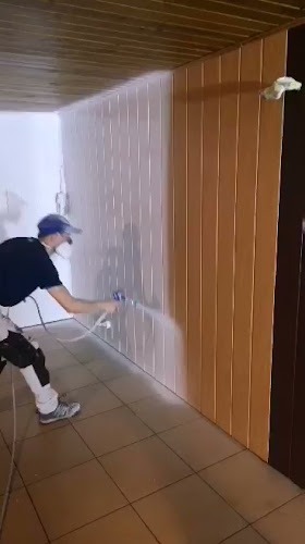 Pracownik w masce maluje ściany w pionowe panele za pomocą pistoletu natryskowego; widoczne zabezpieczenia na kolanach.