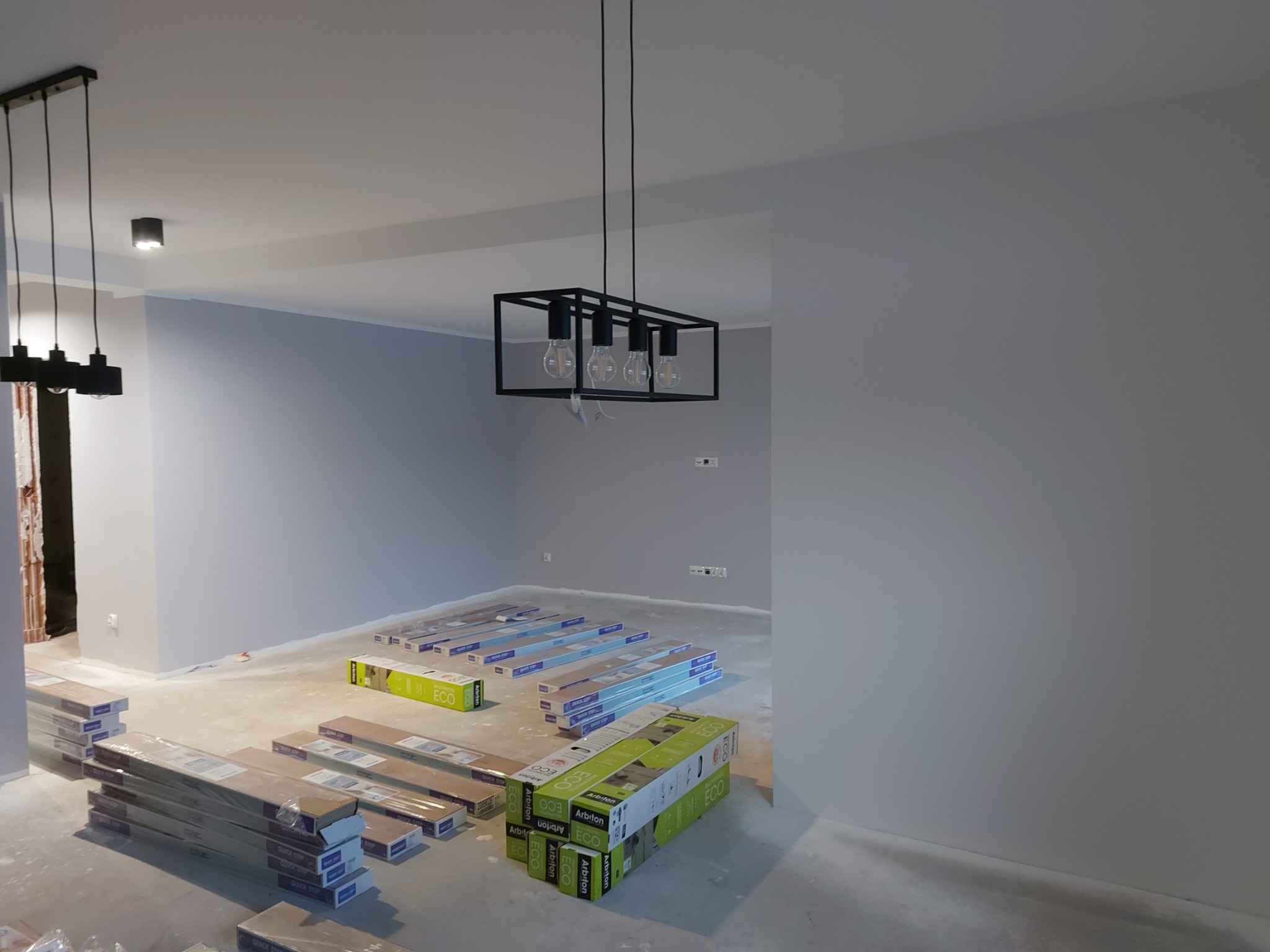 Przygotowanie do położenia paneli podłogowych w nowo pomalowanym pomieszczeniu w Wodzisławiu Śląskim. Widoczne pakiety paneli podłogowych, surowe ściany w odcieniach szarości i nowoczesne lampy...