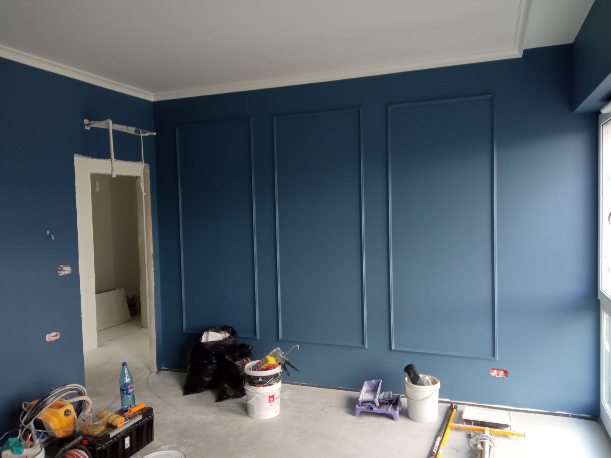 Świeżo pomalowane na granatowo ściany z dekoracyjnymi listwami w pomieszczeniu w trakcie remontu, widoczne narzędzia malarskie i worki na śmieci, gniazdka elektryczne jeszcze nie zamontowane.