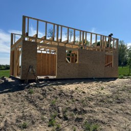 OMT Company sp. z o.o. - Wznoszenie konstrukcji domu z drewna: szkielet z płyt OSB, drabina oparta o ścianę, pracownik na dachu, błękitne niebo w tle. Etap budowy w słoneczny dzień.