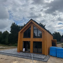 OMT Company sp. z o.o. - Nowoczesny dom szkieletowy z drewnianą elewacją i dużymi oknami, częściowo wykończony taras z kostki brukowej, widoczne materiały budowlane i pracownik przy drzwiach.