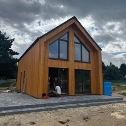 OMT Company sp. z o.o. - Nowoczesny dom z drewna w trakcie wykończenia, z dużymi oknami i tarasem z kostki. Widoczni pracownicy wewnątrz, pochmurne niebo w tle.