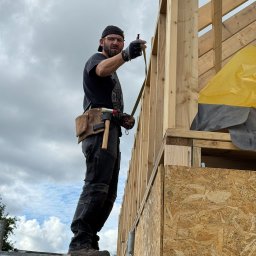 OMT Company sp. z o.o. - Monter w czapce, mierzy konstrukcję domu szkieletowego. Widoczne drewniane elementy, płyty OSB i narzędzia. Ujęcie z dołu, na tle pochmurnego nieba.