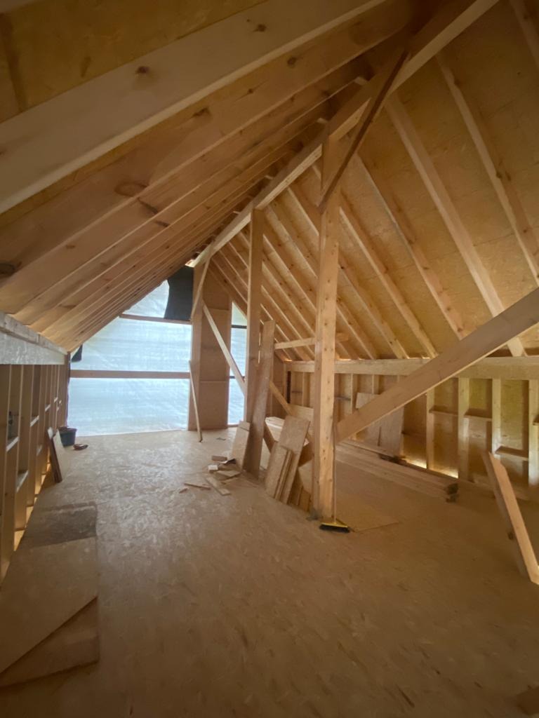 Wnętrze w trakcie budowy, widoczne drewniane elementy konstrukcyjne dachu i ścian, podłoga z płyt OSB, folia budowlana w tle.