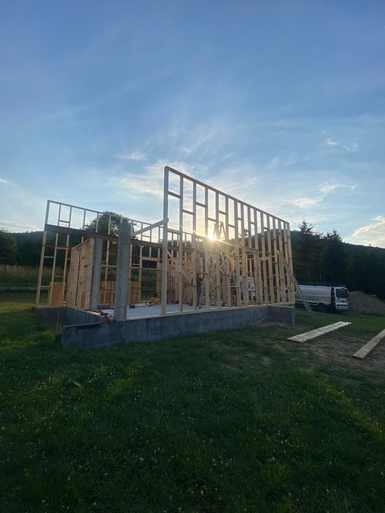 Wznoszenie konstrukcji szkieletowej domu na betonowym fundamencie, widok na szkielet w promieniach zachodzącego słońca, na trawie leżą deski, w tle zaparkowany biały bus.