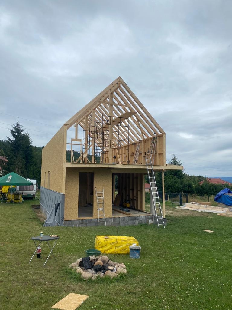 Wznoszenie dwukondygnacyjnej konstrukcji z drewna na fundamencie, widoczny szkielet dachu i ściany z płyt OSB, drabiny opierają się o budynek, palenisko z kamieni na trawie.