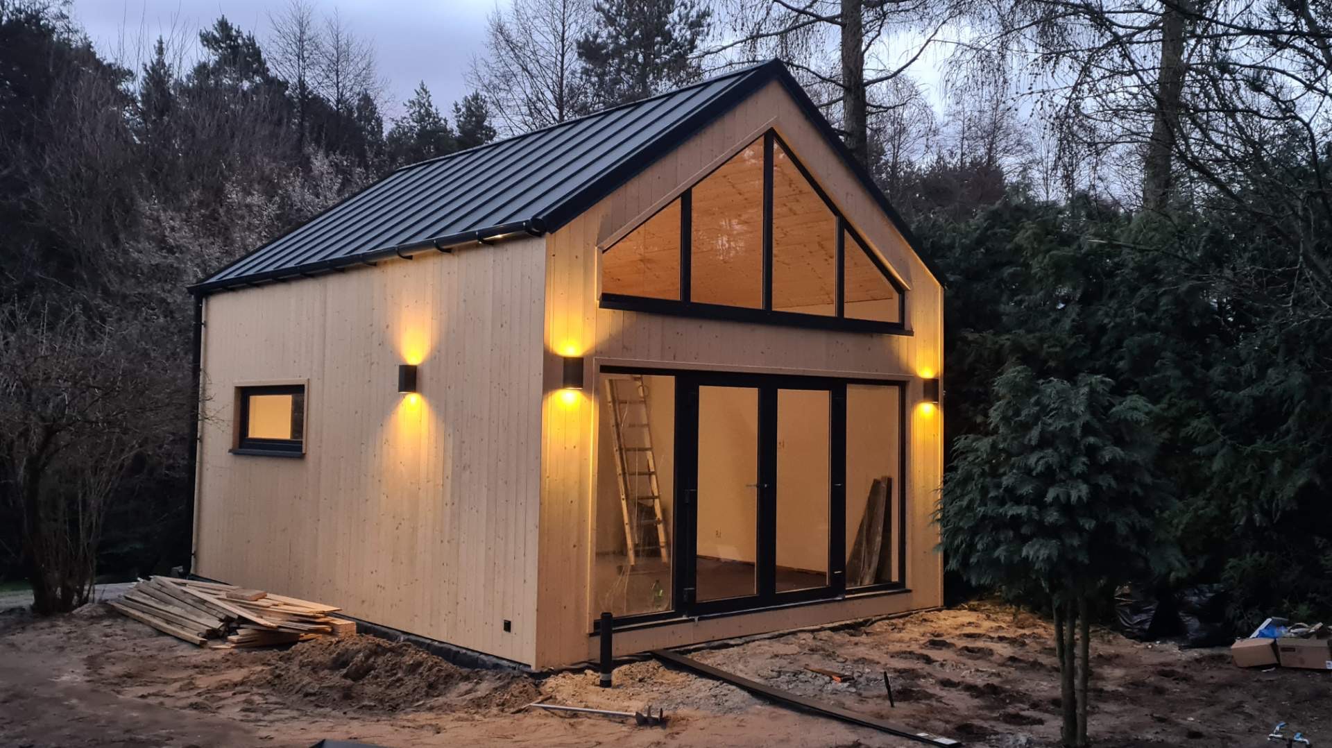 Nowoczesny, surowy dom z drewna z czarną blachodachówką w Luboniu, oświetlony zewnętrznymi lampami, z dużymi przeszkleniami i drabiną opartą o ścianę.