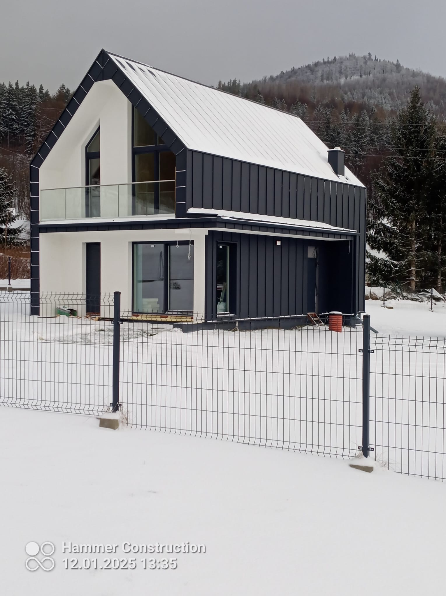 Nowoczesny dom szkieletowy z ciemną elewacją i balkonem ze szklaną balustradą, otoczony zimowym krajobrazem i siatką ogrodzeniową.